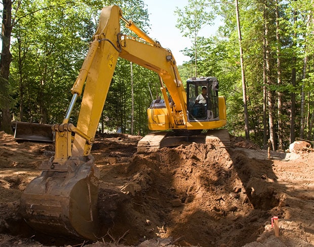 An excavation contractor operating a yellow excavator to dig a hole in the woods.